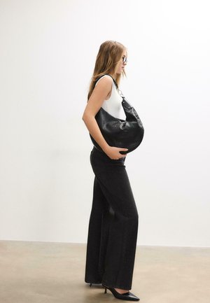 Femme en haut blanc sans manches, jean noir à jambes larges et talons, tenant un grand sac à épaule noir, debout de profil contre un mur blanc.