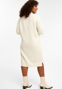 Knielanges cremefarbenes Strickkleid aus weichem Material, mit geripptem Saum, seitlichen Schlitzen und kombiniert mit cremefarbenen Ankle Boots mit Blockabsätzen.