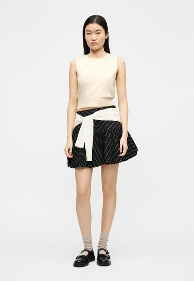 Γυναίκα με μακριά σκουρόχρωμα μαλλιά, που φοράει μπεζ αμάνικη μπλούζα, μαύρη φούστα με ριγέ γραμμές, δεμένο κρεμ πουλόβερ, μπεζ κάλτσες και μαύρα παπούτσια, που στέκεται μπροστά σε λευκό φόντο.