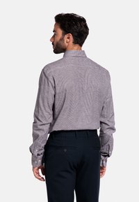 Man met donker haar, gekleed in een geruite blouse met lange mouwen en donkere broeken, staat met de rug naar de camera tegen een effen achtergrond.