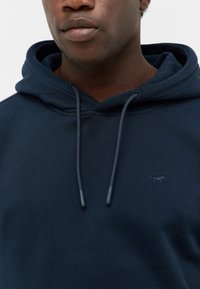 Zbliżenie na osobę ubraną w granatowy hoodie z sznurkami oraz małym, wyszywanym logo konia po lewej stronie klatki piersiowej.