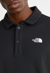 Svart polo-skjorte laget av teksturert stoff, med en to-knapps frontklaff og en hvit logo av "The North Face" på venstre bryst.