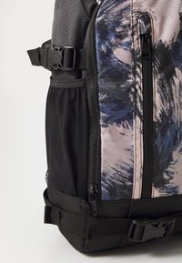 Mochila negra y con estampado que presenta una tela texturizada, múltiples cremalleras y correas ajustables con un bolsillo lateral para botellas.