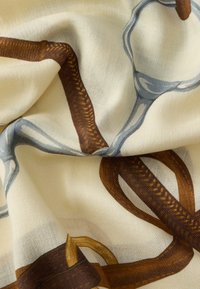 Šál so vzorom s krémovým základom a hnedo-modrými abstraktnými vzormi, hladkou textúrou a jemným leskom. Viditeľný výrazný rybí kostrový vzor.