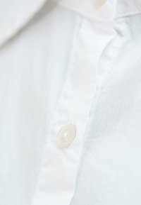 Szczegóły białej koszuli z guzikami na górze, z przezroczystym plastikowym guzikiem, gładką teksturą tkaniny i subtelnym przeszyciem wzdłuż krawędzi.