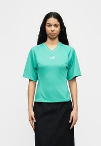 Γυναίκα με μακριά σκούρα μαλλιά που φοράει γαλάζιο αθλητικό μπλουζάκι Puma με V-λαιμόκοψη και μαύρη φούστα, στέκεται μπροστά σε απλό λευκό φόντο.