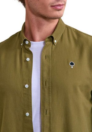 Homme portant une chemise boutonnée vert olive sur un t-shirt blanc à col rond, montrant un logo d'arbre brodé sur la poitrine de la chemise.