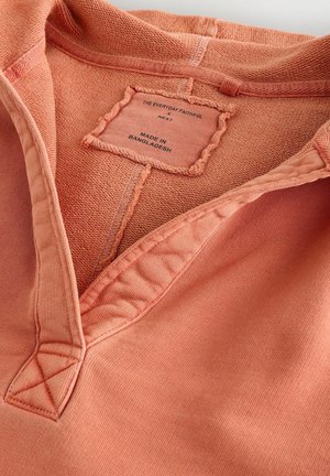 Pull en pêche avec un tissu texturé, présentant un décolleté en V et des détails cousus. L'étiquette indique "Fabriqué au Bangladesh."