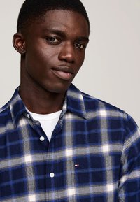 Camicia di flanella a quadretti blu e bianchi con colletto button-down, caratterizzata da un piccolo logo sulla tasca del petto e un tessuto texturizzato.