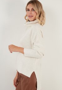 Helle beige Rollkragensweater mit langen Ärmeln, seitlichen Schlitzen und entspanntem Schnitt, vor einem neutralen Hintergrund präsentiert.