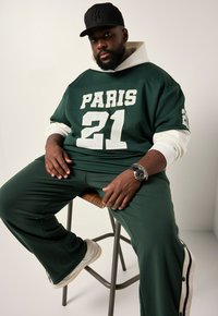 Conjunto atlético verde con "PARIS 21" en malla blanca en la camiseta. Los pantalones cuentan con botones laterales. Combinado con una gorra negra y un reloj plateado.