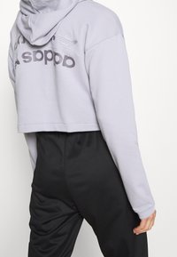 Zkrácená fialová mikina s kapucí, se šedým textem a logem na zadní straně, spárovaná s černými tepláky, ukazující uvolněný fit.