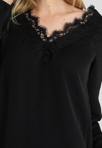 Schwarze Bluse mit langen Ärmeln und V-Ausschnitt, verziert mit aufwendiger schwarzer Spitzenborte. Weiches Material mit glatter Textur.