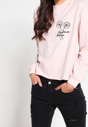 Kobieta w jasnoróżowej, długorękawowej koszuli z napisem „Daydream Believer” i motywem mniszka lekarskiego, w czarnych podartych dżinsach, z rękami w kieszeniach.