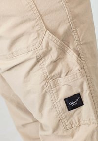 Nahaufnahme von beigen Stoffhosen, die detaillierte Nähte und ein kleines schwarzes, rechteckiges Etikett mit weißer Schreibschrift "Reell est 1997" zeigen.