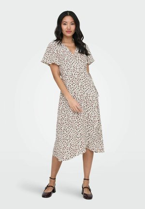 Kvinde iført en hvid midi-kjole med rød blomsterprint, flagrende ærmer, V-udskæring og sorte hæle med ankelrem, stående mod en ensfarvet baggrund.