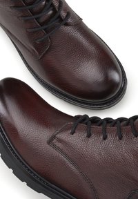 Stivaletti in pelle marrone con finitura texture, punta tonda e lacci neri. Presentano una suola nera robusta e dettagli di cucitura minimi.