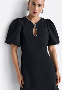 Schwarzes Kleid mit Puffärmeln, ausgestattet mit einem Schlüssellochausschnitt, der mit Kristallverzierungen geschmückt ist. Glatte Textur und figurbetonter Schnitt.