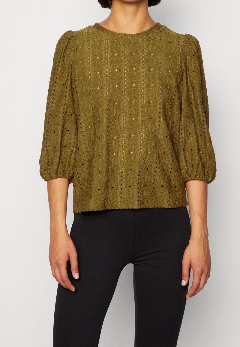 Femme portant un chemisier en maille texturée vert olive avec des manches bouffantes trois-quarts et un pantalon noir ajusté sur un fond uni.