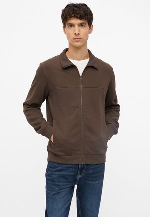 Giovane uomo con capelli scuri e ricci che indossa una giacca marrone con cerniera sopra una camicia bianca e jeans blu, in piedi con le mani in tasca.