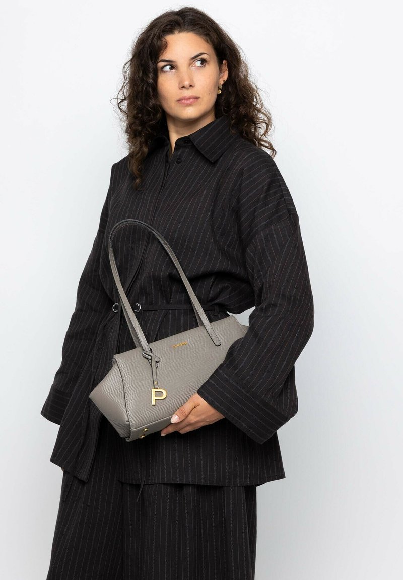 Sac gris avec une texture nervurée, doté d'un charme en lettres dorées et d'une courte poignée, tenu contre une tenue à rayures noires.