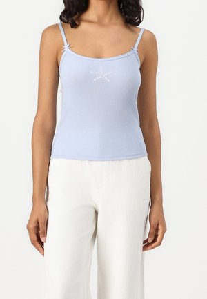 Femme portant un haut bleu clair à fine bretelle avec un motif d'étoile de mer blanche et un pantalon blanc, debout devant un fond uni.