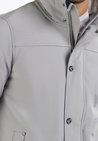 Veste gris clair en tissu lisse, dotée d'une fermeture éclair à l'avant, de boutons-pression, d'une capuche et d'un logo sur le côté gauche.