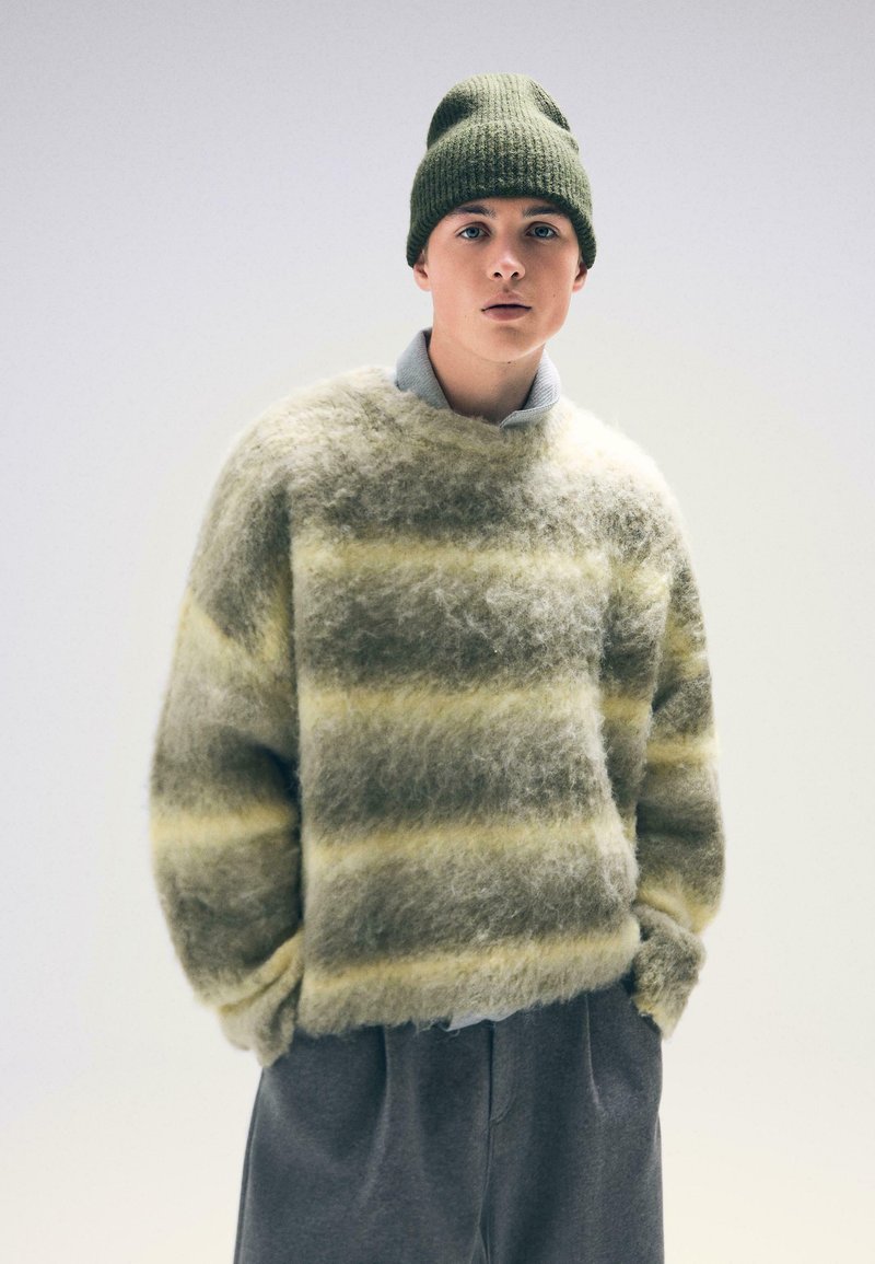 Futrzasty, w paski sweter w zielono-żółtych kolorach, połączony z prążkowaną zieloną czapką. Sweter ma okrągły dekolt i luźny krój.