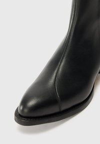 Botín de cuero negro con punta puntiaguda, textura suave, detalle sutil de costuras a lo largo del lateral y una suela delgada de goma.