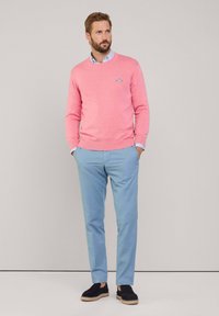 Maglione rosa chiaro, a maniche lunghe, con logo; pantaloni azzurri, fit dritto; scarpe nere con suola intrecciata; sfondo neutro.