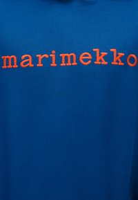 Μπλε βαμβακερό πουκάμισο με πορτοκαλί κεντητό κείμενο που γράφει "marimekko". Λεία υφή με κανονική εφαρμογή και μίνιμαλ σχεδιασμό.