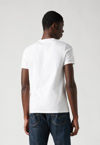 Witte katoenen t-shirt met een klassieke pasvorm en korte mouwen, gedragen door iemand in donkerblauwe jeans met zichtbare achterzakken.