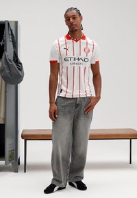 Camisola de futebol branca com riscas vermelhas e detalhes a preto, com o logótipo "ETIHAD AIRWAYS". Combinada com jeans cinzentos largos e sapatos pretos.