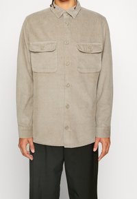 Beige kordfløjlsjakke med krave med knapper, knapper foran, to brystlommer og ribbet tekstur. Lange ærmer med knapmanchetter.