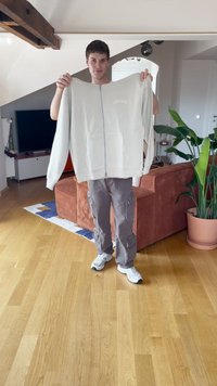 Jaunas vyras stovi patalpose, laiko baltą užtrauktuką su džemperiu, o fone matyti rudas sofą ir žalia augalą.