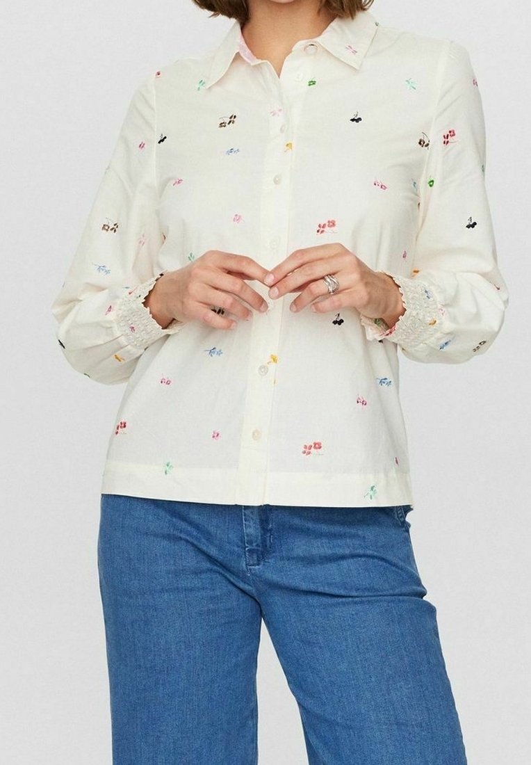 Camicia button-up color crema chiaro con ricami floreali in vari colori. Presenta un colletto classico e polsini elasticizzati. Abbinata a jeans blu.