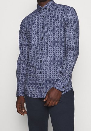 Mann trägt ein eng anliegendes, langärmeliges Hemd mit blauem geometrischem Muster und dunkle Hose, steht vor einfachem Hintergrund.