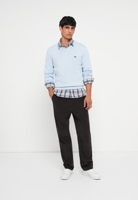 Lichtblauwe v-halstrui over een geruit shirt met blauwe, witte en groene patronen; gecombineerd met donkere broek en witte sneakers.