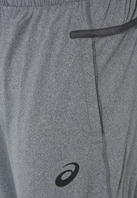 Šedé sportovní kalhoty vyrobené z texturované tkaniny s černým logem. Mají elastický pas a boční kapsy s černými akcenty.