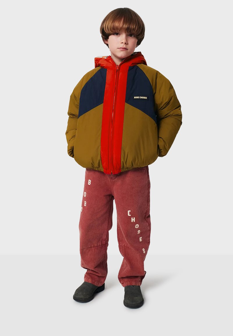 Chaqueta de color verde oliva y azul marino con detalles en rojo, cierre de cremallera y logotipo. Combinada con pantalones de pana rosa con texto blanco. Zapatos negros.