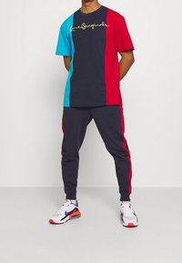 Tričoko s krátkým rukávem v barevném bloku, obsahující námořnickou modrou, červenou a tyrkysovou barvu s žlutým textem, v kombinaci s námořnickými tepláky a vícebarevnými teniskami.