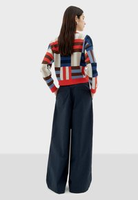 Pull en tricot multicolore à motifs avec des carrés rouges, bleus et bruns ; associé à un pantalon large sombre. Texture lisse et coupe décontractée.