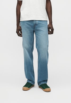 Μαύρο άτομο που φοράει ανοιχτό μπλε τζιν με ίσια μπατζάκια, πράσινα αθλητικά παπούτσια με ταμπά σόλες και άσπρο πουκάμισο, στέκεται μπροστά σε απλό φόντο.