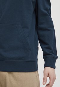 Temno modra kratka majica s kenguru žepom, ribastimi manšetami in robom. Mehka tekstura, okrašena z znamko na spodnji strani.