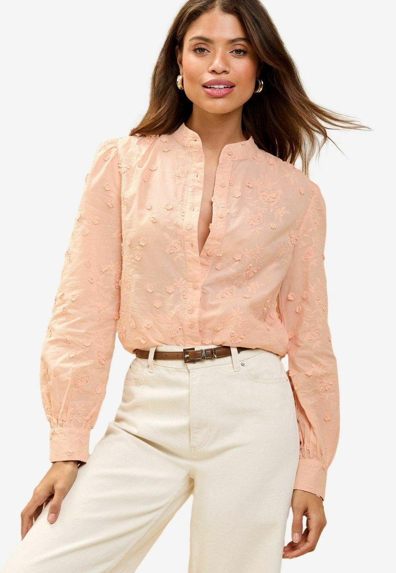 Blusa ricamata pesca con scollatura a collare e maniche a sbuffo, abbinata a pantaloni color crema e una cintura marrone con fibbia argentata.