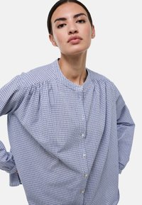 Chemise boutonn�ée à carreaux bleus et blancs avec un col rond, des manches bouffantes et une coupe ample, ornée de petits boutons sur le devant.
