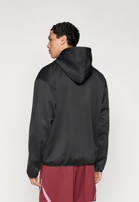 Sweat-shirt à capuche noir avec une texture lisse, doté d'une poche kangourou et de poignets élastiques, associé à un short bordeaux.