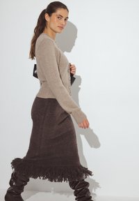 Beežist kootud kardigan nööpidega, kombineeritud tumepruuni fringe-seeliku ga. Modell kannab läikivaid musti põlveni ulatuvaid saapaid.
