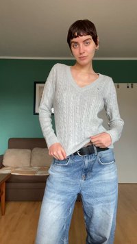 Szary sweter w splot kabelkowy z dekoltem w serek, zestawiony z jasnoniebieskimi jeansami o szerokich nogawkach. Sweter ma ściągacze przy mankietach oraz czarny pasek.