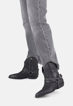 Personne portant un jean gris et des bottines en cuir noir avec fermetures éclair latérales, sangles décoratives et talons bas sur un fond clair.